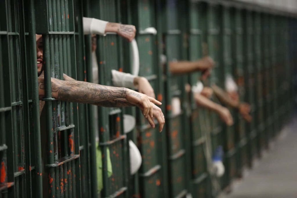 Gritty prison wallpaper showing tattooed inmates reaching through dark green cell bars corridor
