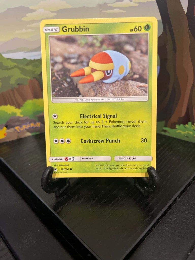 Grass-type Grubbin Pokémon card displayed on black stand with forest art backdrop by Yuka Morii Wallpaper