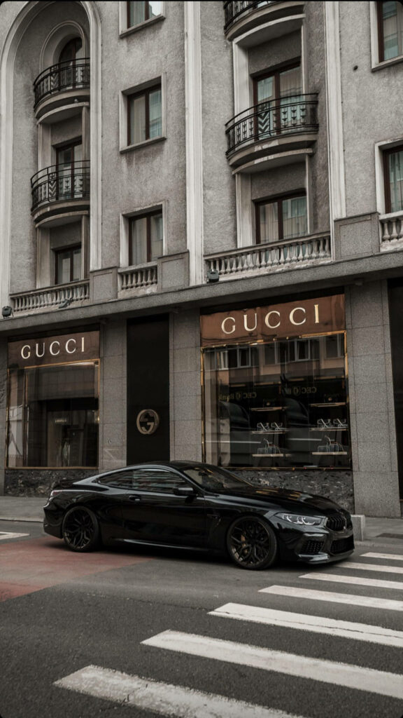 Luxury Defined: A Gucci Storefront Enveloped by Glamour, with a Sleek Black Car Completing the Scene - Custom Gucci iPhone Wallpaper