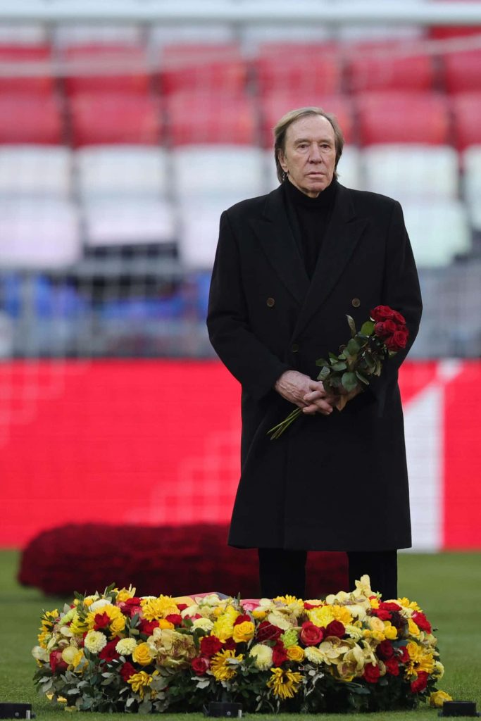 Günter Netzer tribute in stadium holding red roses near colorful floral wreath with red seats Wallpaper