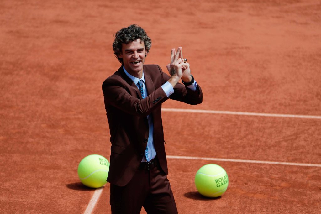 Gustavo Kuerten smiling on Roland Garros clay court in brown suit with giant tennis balls background Wallpaper