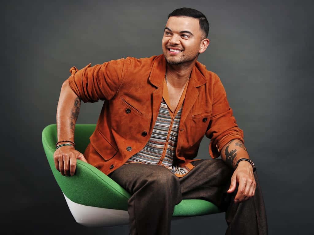 Guy Sebastian stylish portrait rust jacket striped shirt seated green chair neutral backdrop Wallpaper
