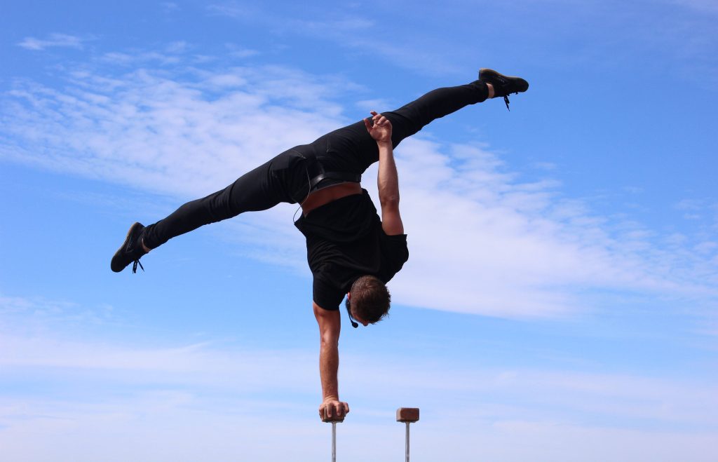 Male gymnast in one-arm handstand split on canes against bright cloudy blue sky wallpaper
