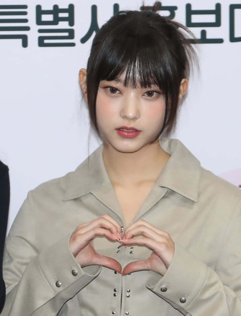Haerin from NewJeans posing with finger heart in beige studded jacket wallpaper background