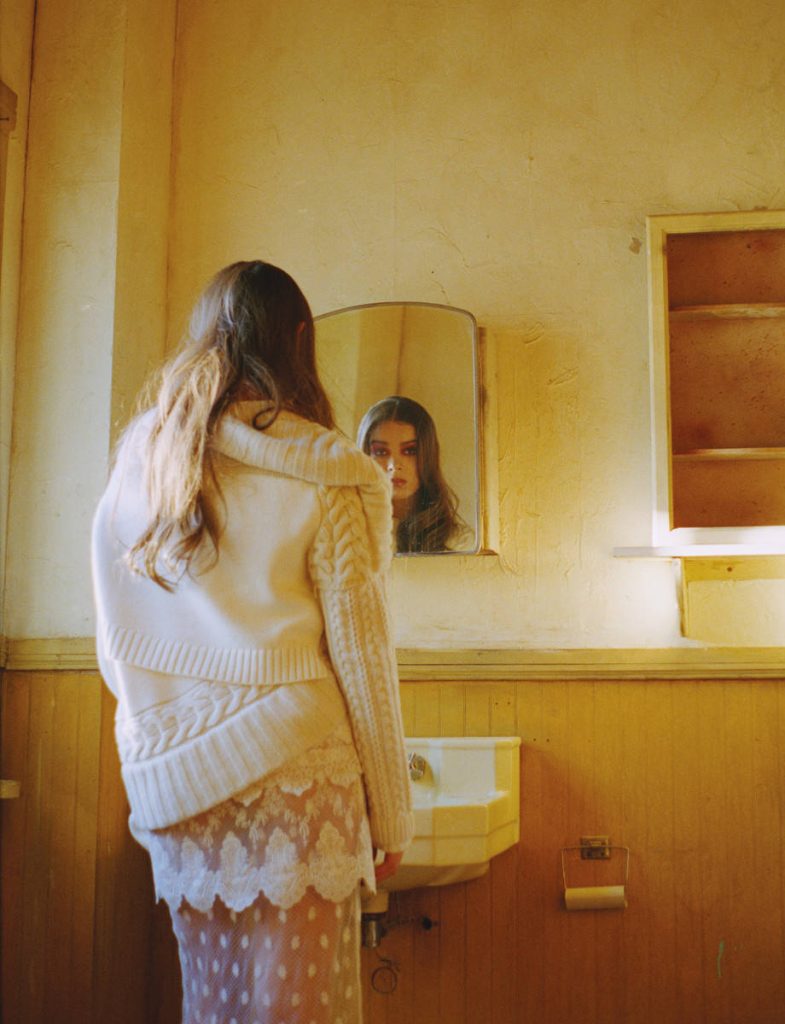 Hailee Steinfeld in cozy cable-knit sweater and lace skirt gazing into mirror vintage room Wallpaper