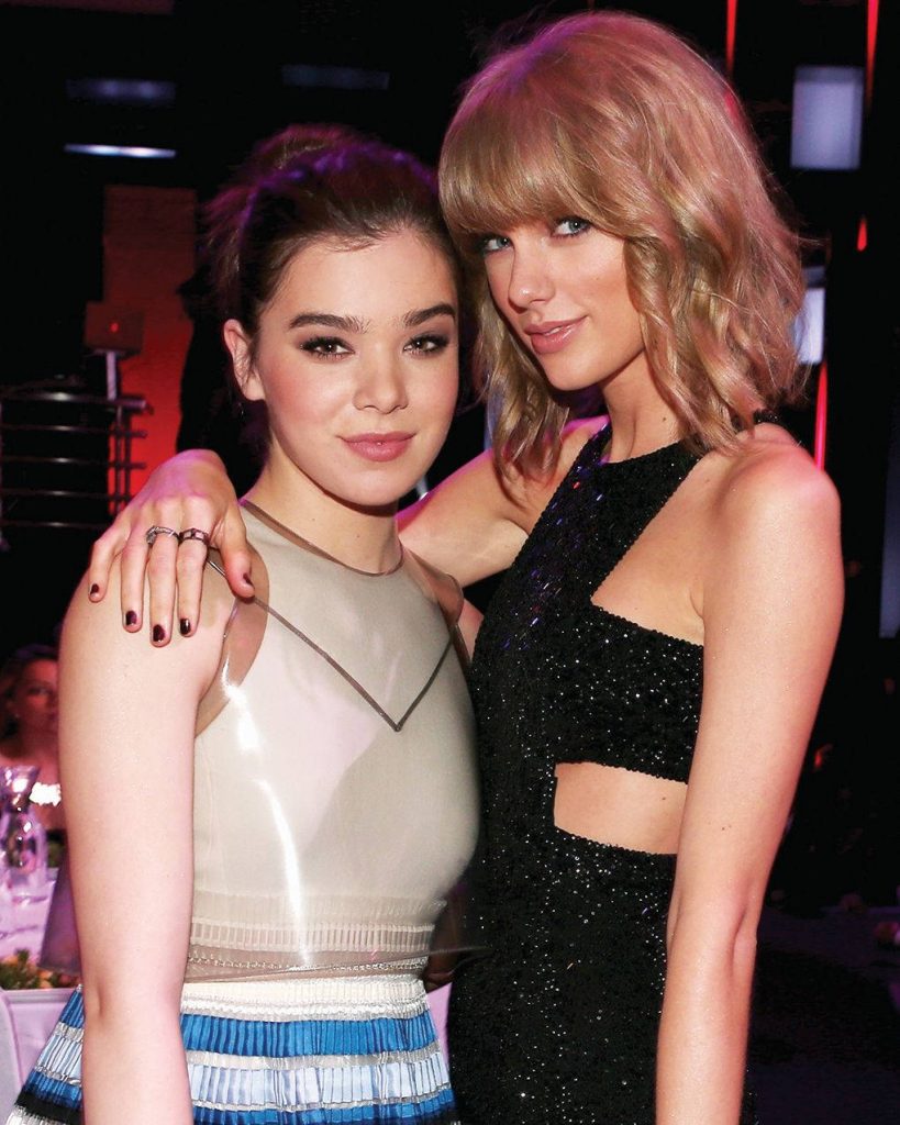Hailee Steinfeld and Taylor Swift posing at glamorous event in elegant outfits and red lighting Wallpaper