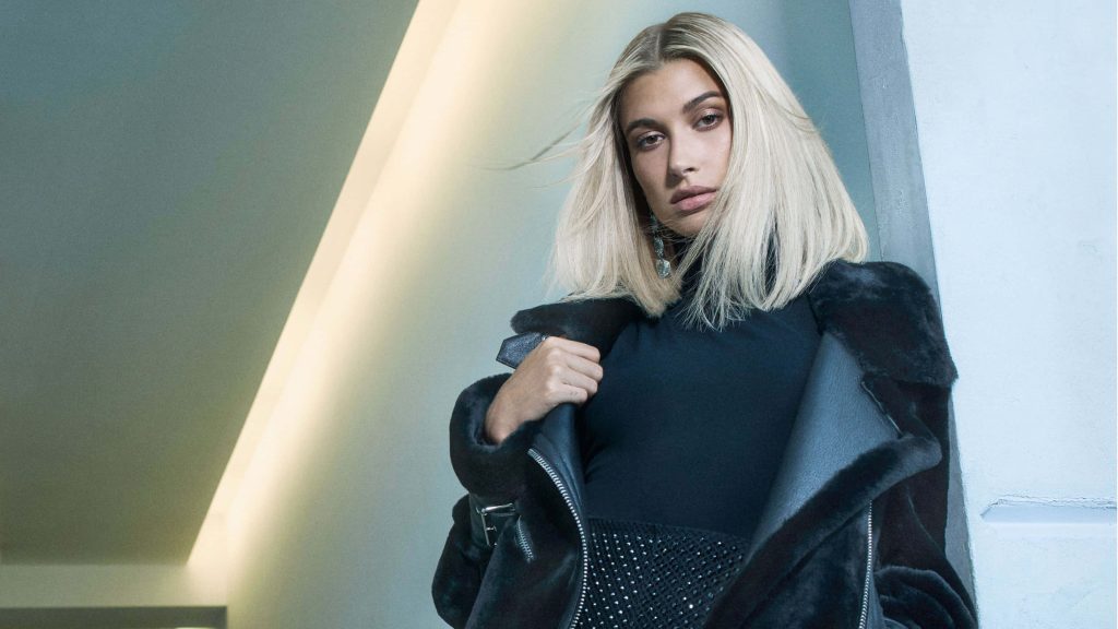 Hailey Bieber in black high-neck top and fur jacket against minimalist cool-toned backdrop wallpaper