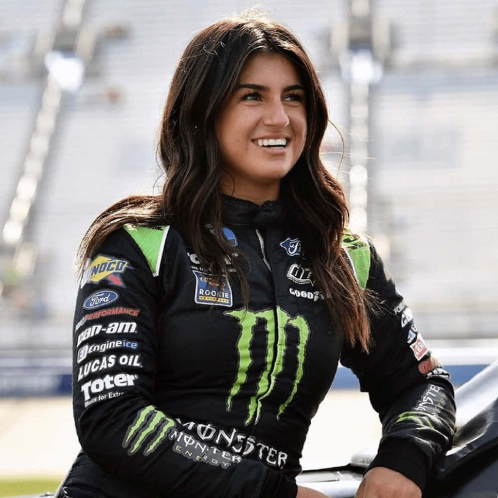 Hailie Deegan in black Monster Energy suit smiling on race track with blurred grandstand background Wallpaper