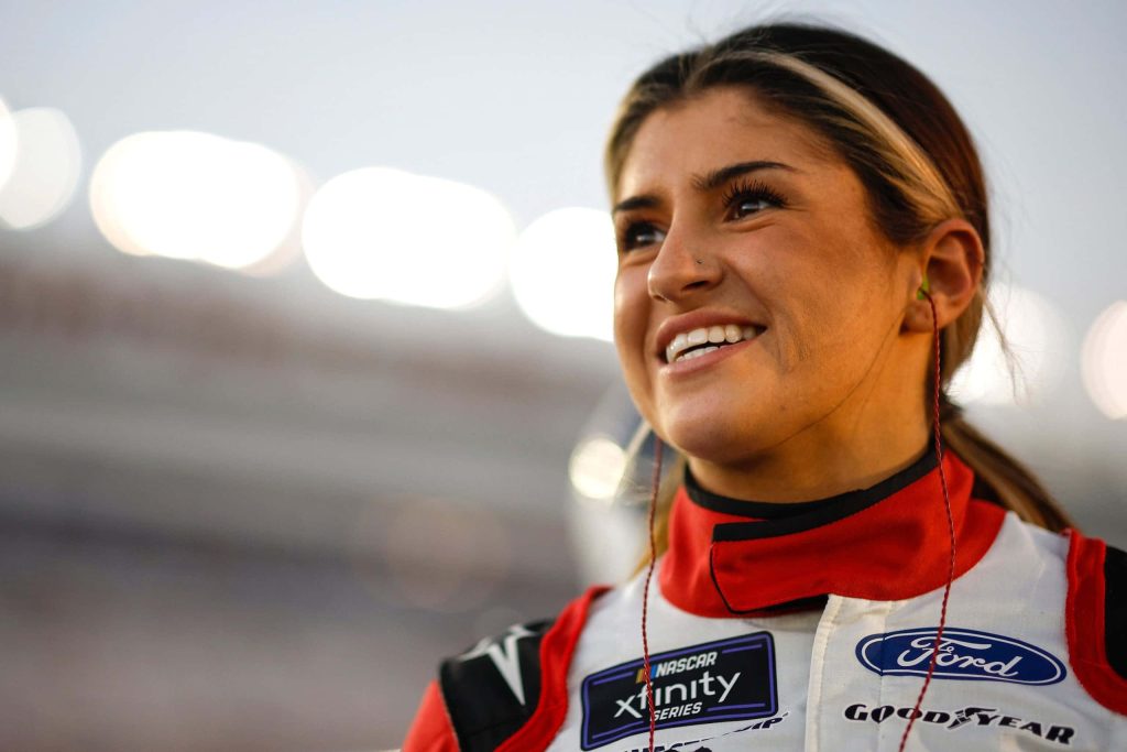 Hailie Deegan smiling in racing suit soft-lit portrait blurred stadium vibrant NASCAR wallpaper