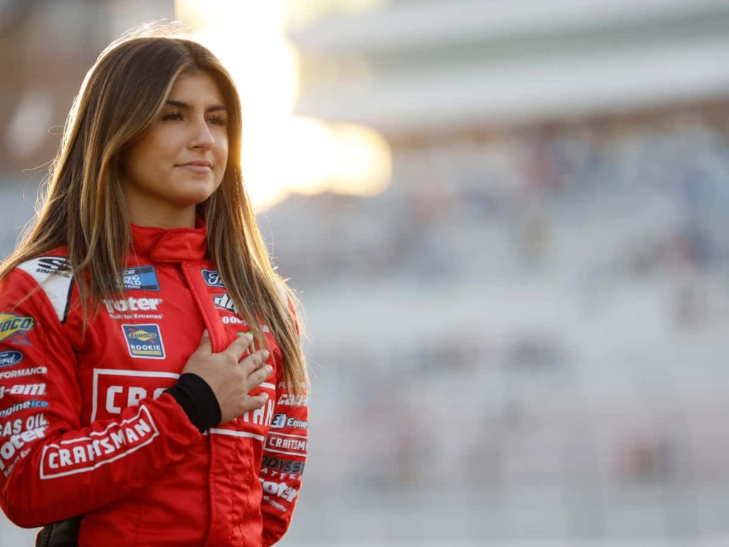 Hailie Deegan in red NASCAR suit hand over heart portrait with blurred racetrack background Wallpaper