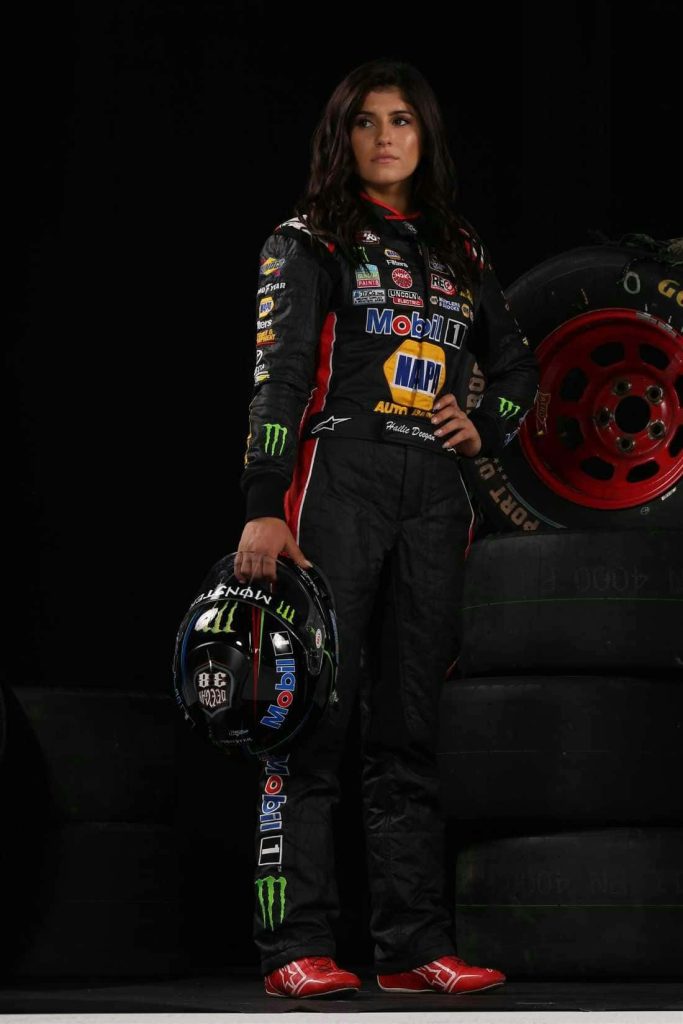 Racing driver Hailie Deegan in black red firesuit holding helmet beside tires studio wallpaper background