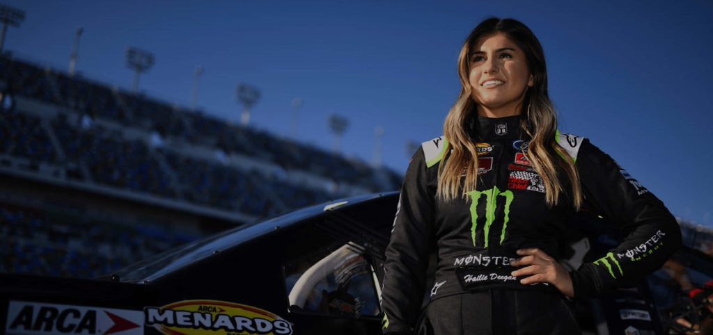 Hailie Deegan in black Monster Energy suit standing trackside with blurred race track background Wallpaper
