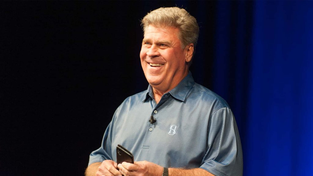 Golfer Hal Sutton in blue polo smiling holding smartphone on stage with blue lighting background Wallpaper