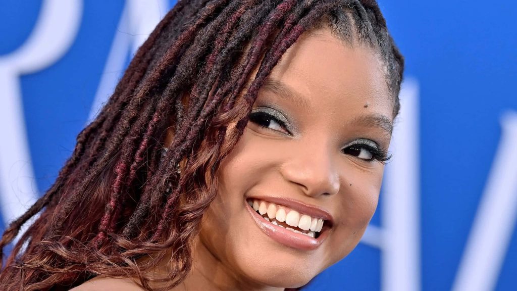 Halle Bailey smiling close-up with red locs and shimmering makeup on vivid blue background wallpaper