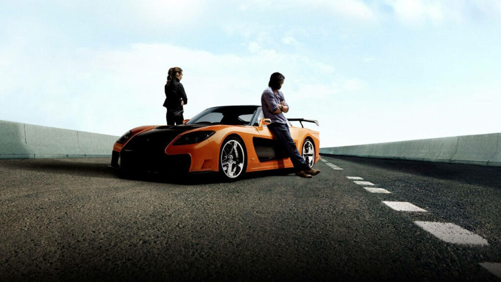 Han Lue leans on orange car on open road clear sky Fast and Furious cool confident wallpaper