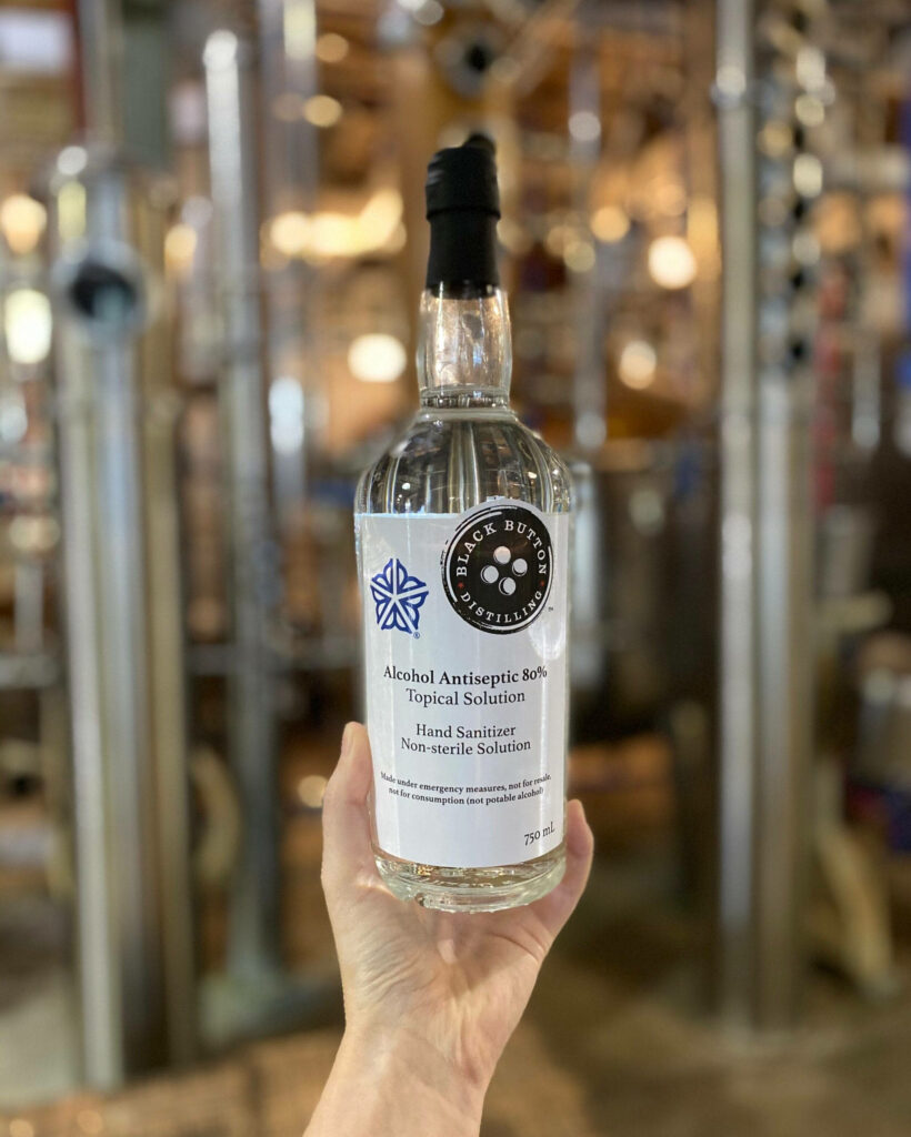 Hand holding Black Button Distilling alcohol hand sanitizer bottle in distillery with metal equipment Wallpaper