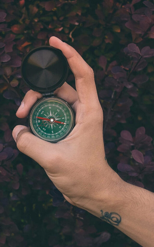 Hand holding open compass with wrist tattoo against deep purple foliage adventure wallpaper background