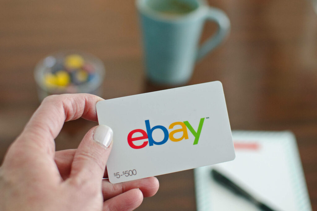 Hand holding eBay gift card with multicolored logo near blue coffee mug in modern workspace Wallpaper
