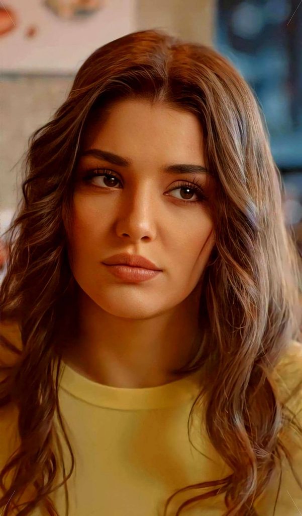 Hande Erçel close-up portrait with long wavy hair in yellow top warm-toned wallpaper background