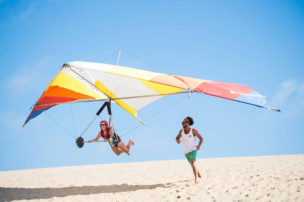 Woman hang gliding over golden dunes at Jockey's Ridge State Park under clear blue sky Wallpaper