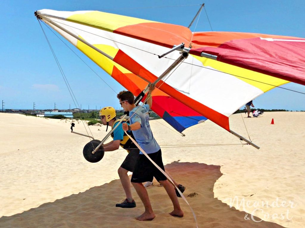 Hang gliding lesson on sunny sand dune with instructor guiding student in yellow helmet flying Kitty Hawk Wallpaper