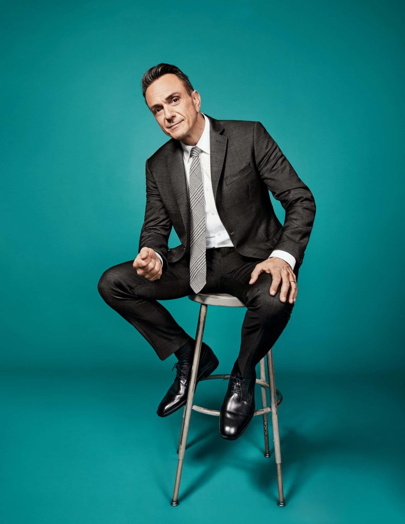 Hank Azaria in charcoal grey suit seated on metal stool with teal studio background wallpaper