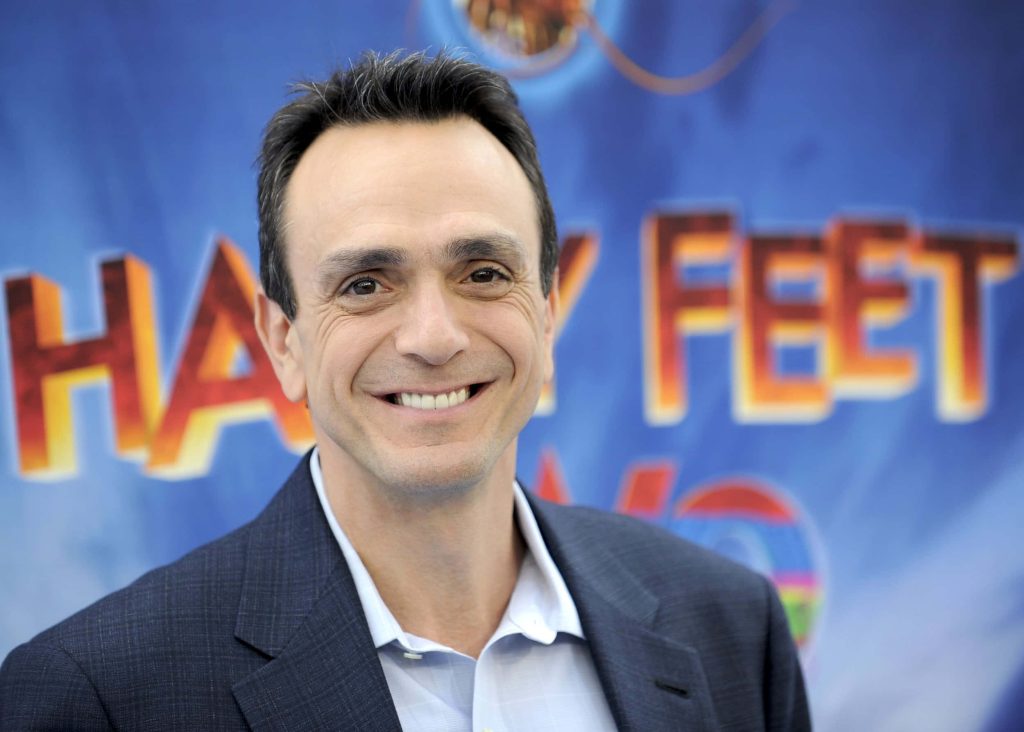 Hank Azaria smiling in blue blazer with vibrant colorful Happy Feet movie backdrop wallpaper