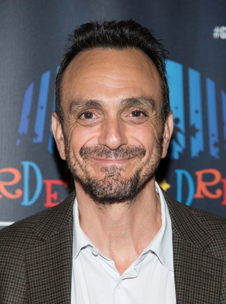 Hank Azaria smiling in casual blazer at press event with dark branded colorful backdrop wallpaper