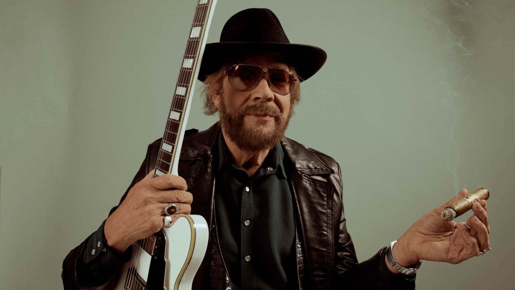 Hank Williams Jr in leather jacket with white electric guitar and cigar against green backdrop Wallpaper