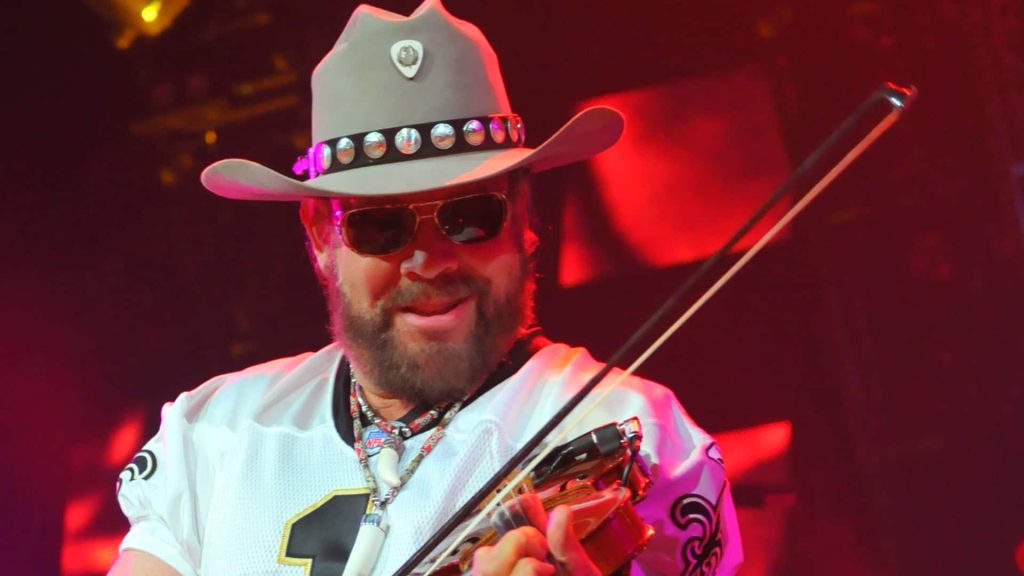 Hank Williams Jr playing fiddle live in New Orleans Saints jersey and cowboy hat red stage lighting Wallpaper