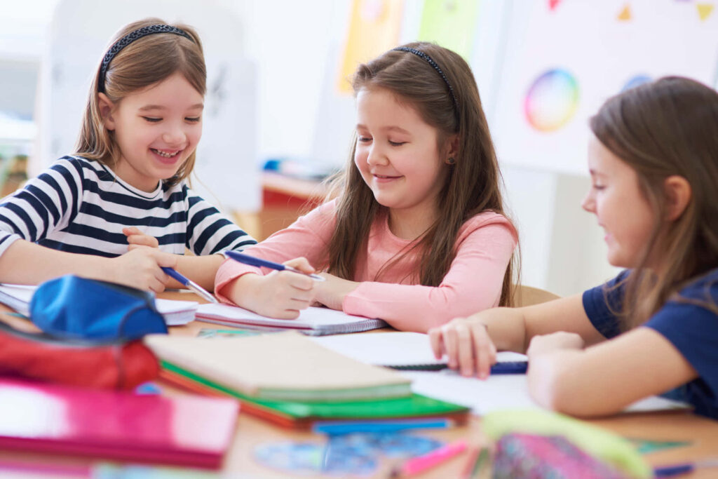 Three young girls smiling and collaborating at a colorful classroom table with educational decor Wallpaper