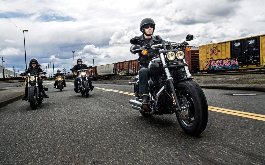 Harley-Davidson FXDF Fat Bob riders cruising past industrial railcars under cloudy sky wallpaper