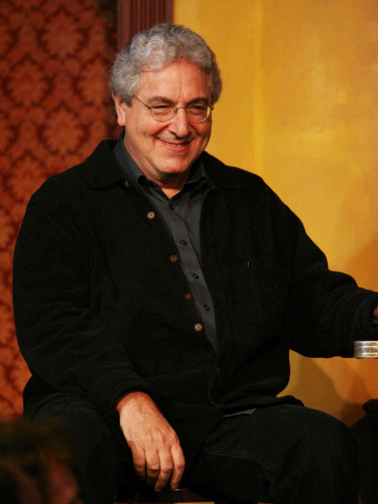 Portrait of filmmaker Harold Ramis smiling in black jacket glasses warm yellow patterned background Wallpaper