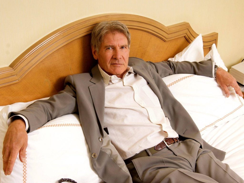 Harrison Ford in tailored grey suit sitting relaxed against wooden headboard with soft lighting Wallpaper