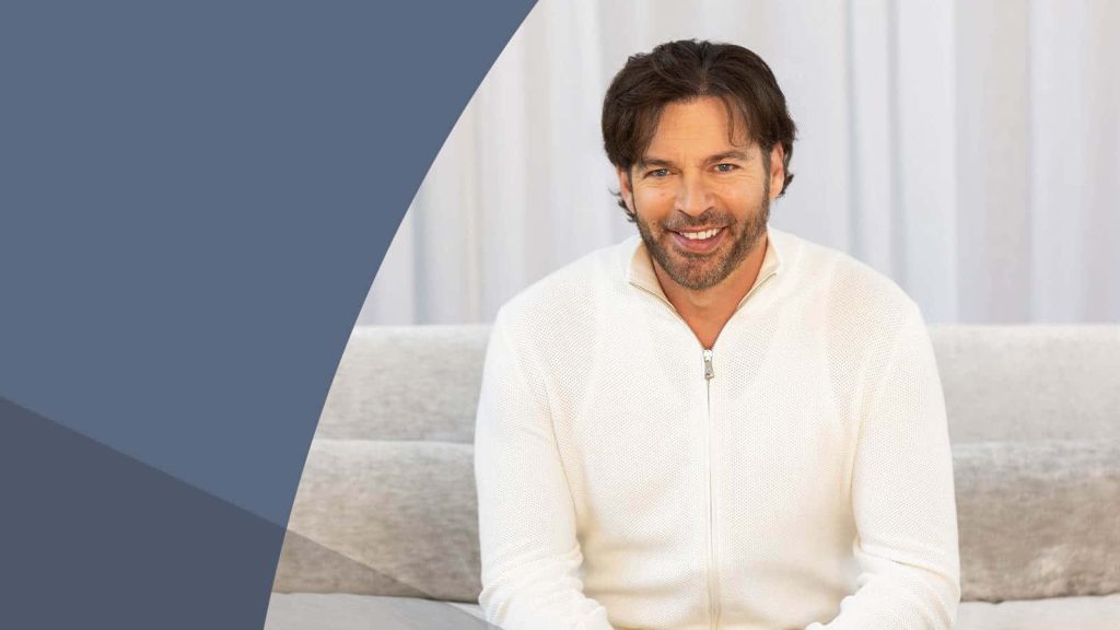 Harry Connick Jr. smiling in cream sweater on light couch with blue geometric background wallpaper