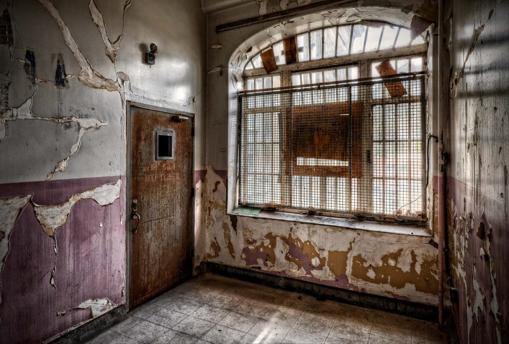 Decaying prison interior with rusted fire door and barred window in abandoned jail wallpaper