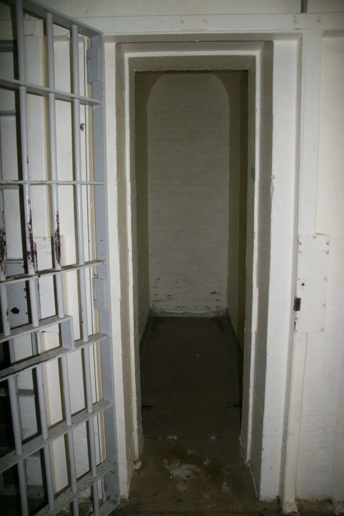 Open barred metal jail door revealing dimly lit prison cell with white masonry walls background Wallpaper