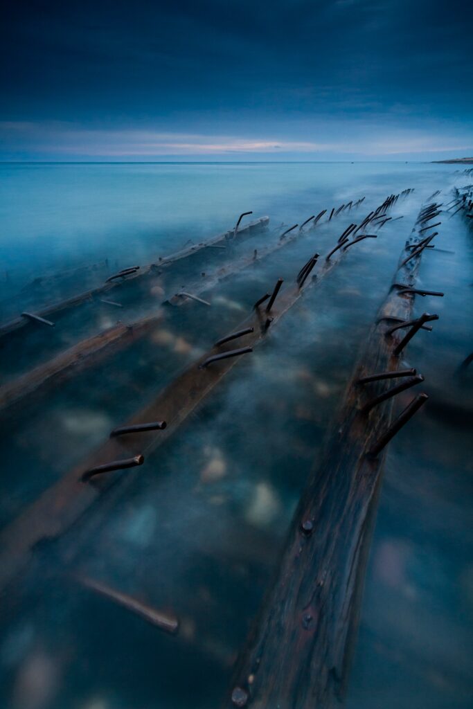 Haunting shipwreck with weathered wood and rusted spikes in misty blue water under moody sky Wallpaper
