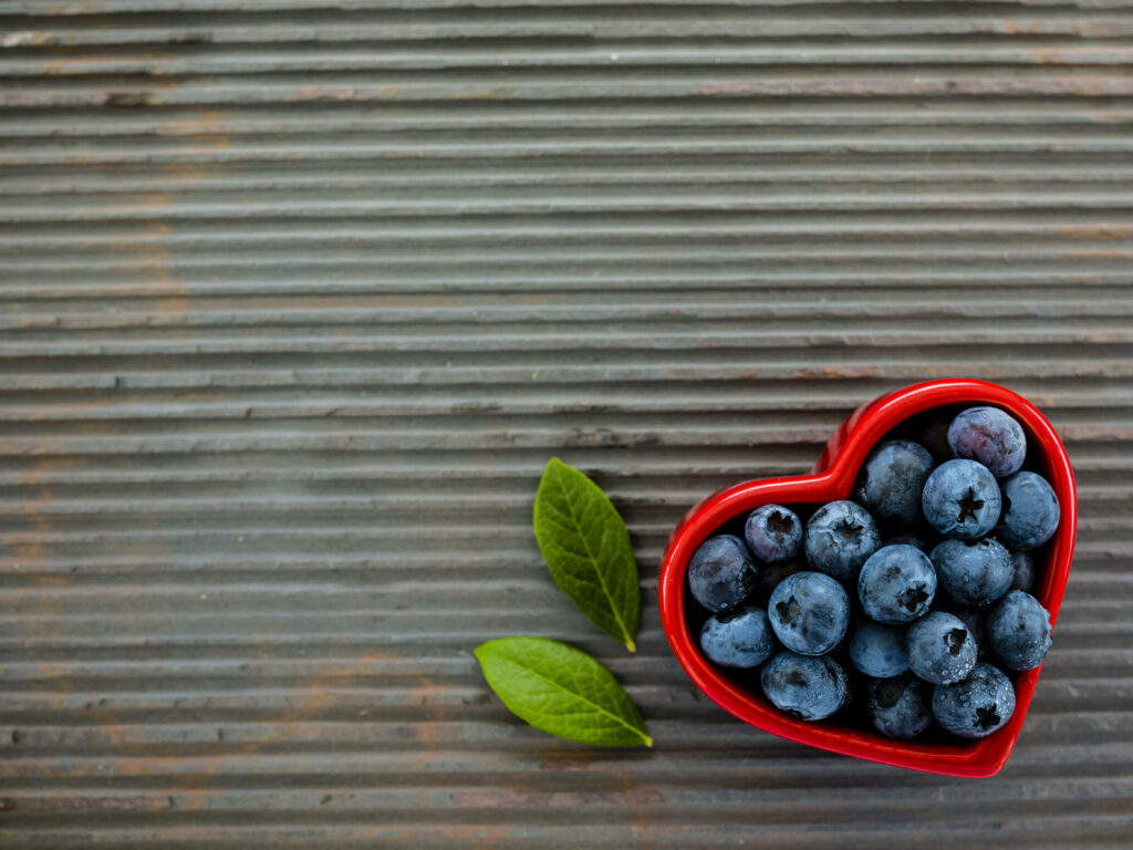 Heart-shaped ramekin filled with fresh blueberries on wooden surface with green leaves background Wallpaper