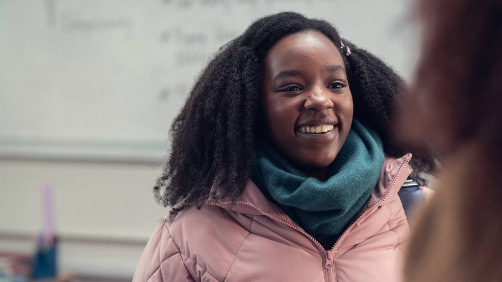 Elle Argent Heartstopper smiling warmly in pink jacket and teal scarf classroom wallpaper background