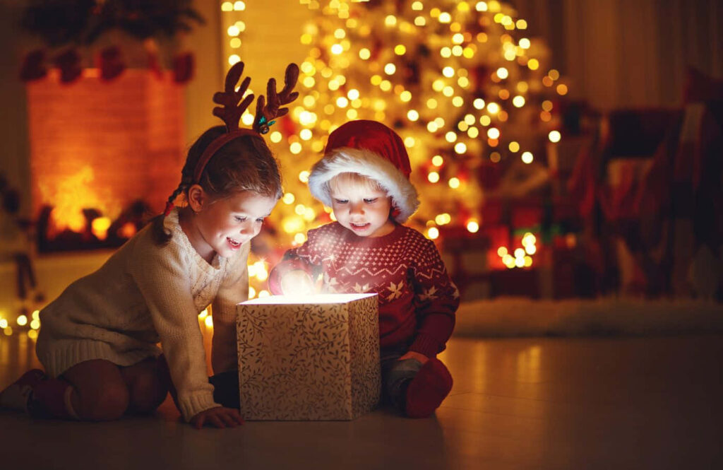 Joyful Kids Unwrap Gift in Cozy Holiday Scene with Christmas Tree Wallpaper