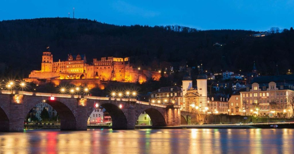 Heidelberg Castle glowing at night with Old Bridge reflected in Neckar River under blue sky Wallpaper