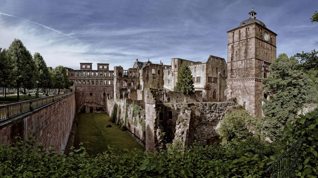 Heidelberg Castle ruins with red sandstone clock tower surrounded by greenery under cloudy sky Wallpaper