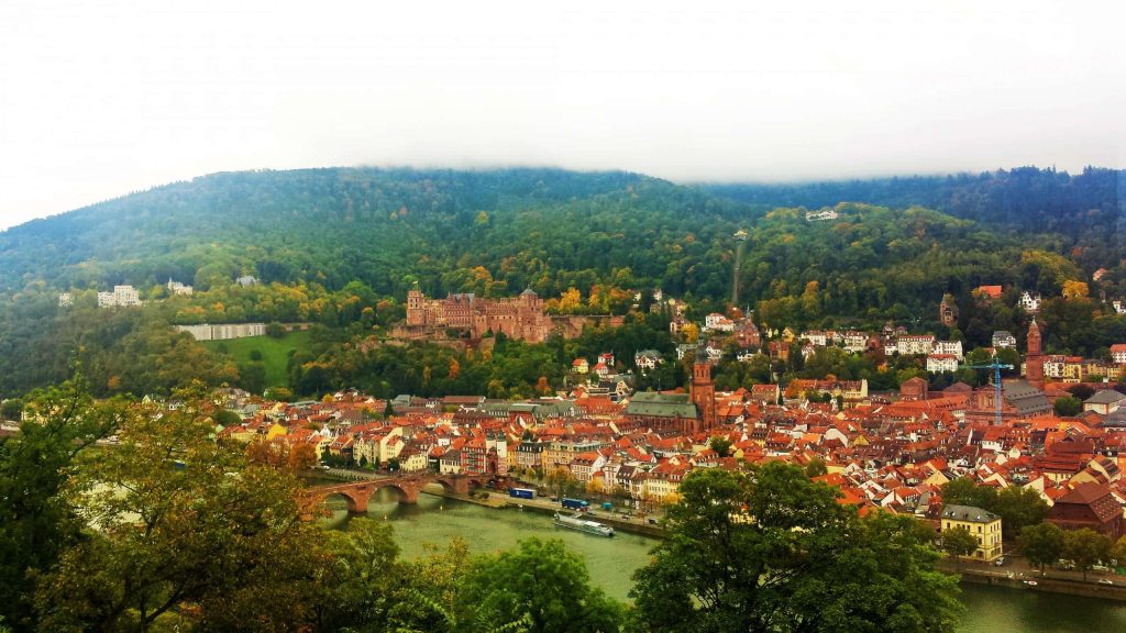 Heidelberg Castle on forested hill under cloudy sky with red rooftops and Neckar River view Wallpaper