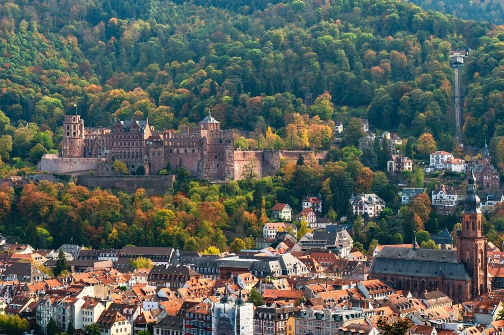 Aerial view of historic Heidelberg Castle with autumn forests and red-roofed cityscape below Wallpaper