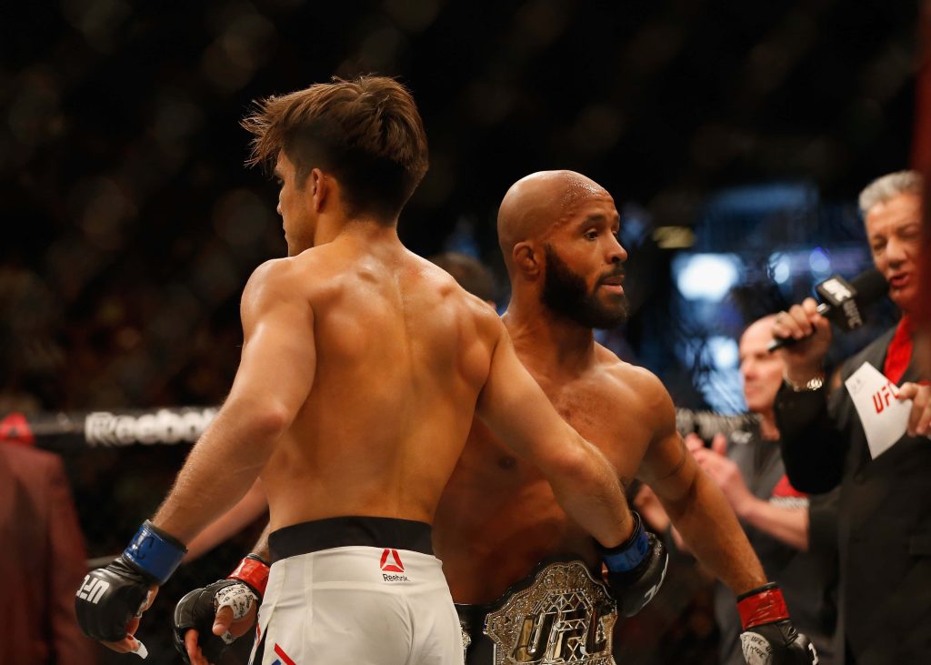 UFC fighters Henry Cejudo and Demetrious Johnson post-match in intense Octagon with championship belt Wallpaper