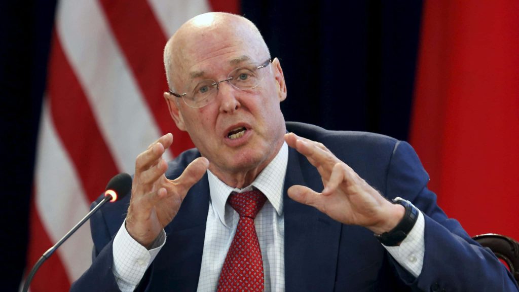 Henry Paulson speaking at US China summit in dark suit and red tie with national flags background Wallpaper
