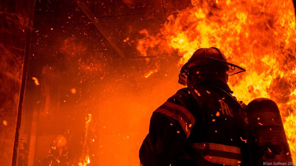 Firefighter in full gear battles blazing orange fire with flying embers heroic action scene Wallpaper