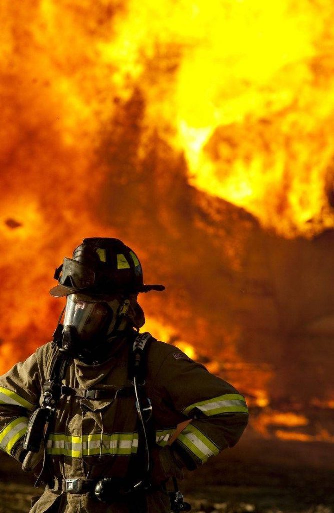 Heroic firefighter in full gear with SCBA mask stands before intense blazing fire background wallpaper