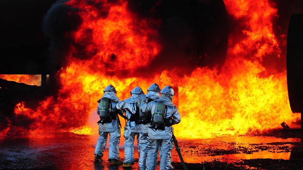 Four firefighters in aluminized suits with MSA gear face blazing fire wallpaper background